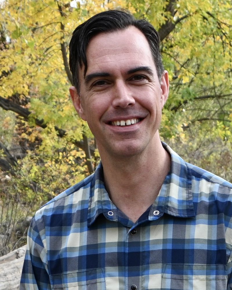 David smiling in a plaid shirt among golden autumn trees