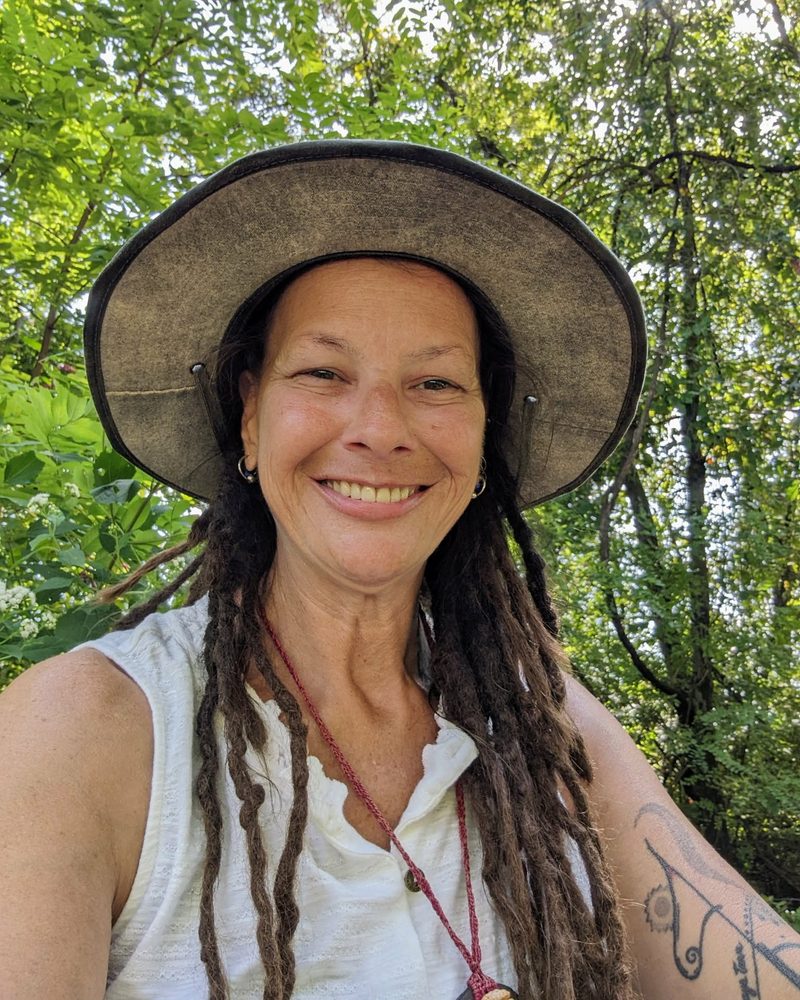 Lynn smiling in a wide-brimmed hat among green trees